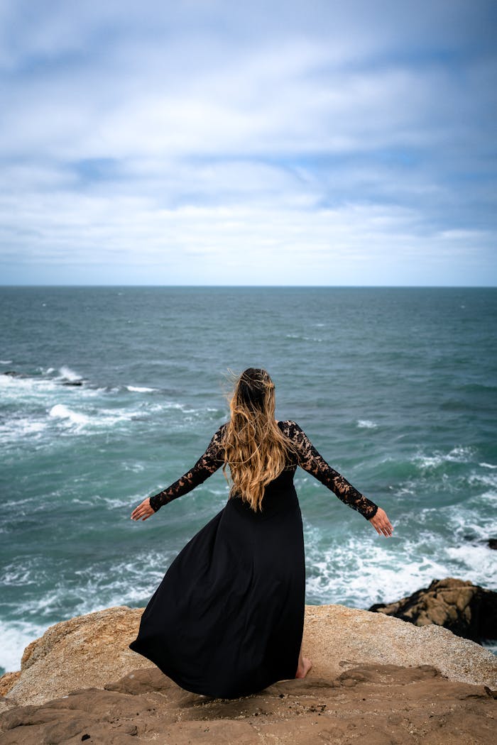 A woman in a black dress with long hair stands on rocks overlooking the ocean, embodying elegance and freedom.