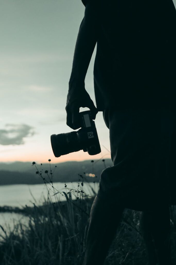 A photographer holding a camera in silhouette against a serene sunset backdrop.