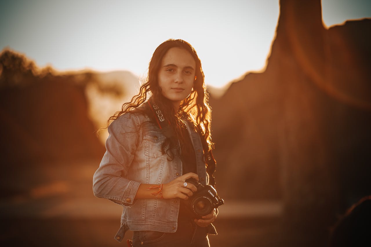 Pretty female with brown curly hair holding camera and standing under bright sunlight while looking at camera